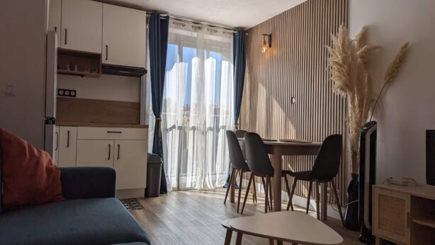 Een moderne woonkamer met een bank, een eethoek met stoelen, een keuken met keukenkastjes en een groot raam met gordijnen.
