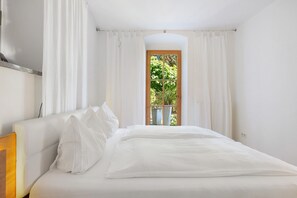 Una camera da letto con un letto, tende bianche, una porta in legno e vista sul verde.