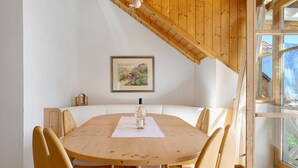 Ein Essbereich mit einem Holztisch, weißen gepolsterten Stühlen, einem gerahmten Bild an der Wand und einem Fenster mit Blick ins Freie.