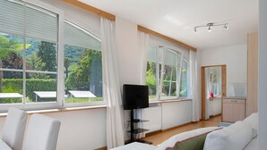 Et værelse med en hvid sofa, et fjernsyn på et stativ og store vinduer med gardiner.