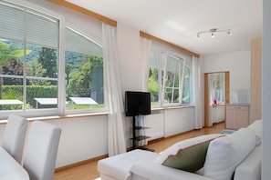 Um quarto com sofá branco, televisão em móvel e amplas janelas com cortinas.