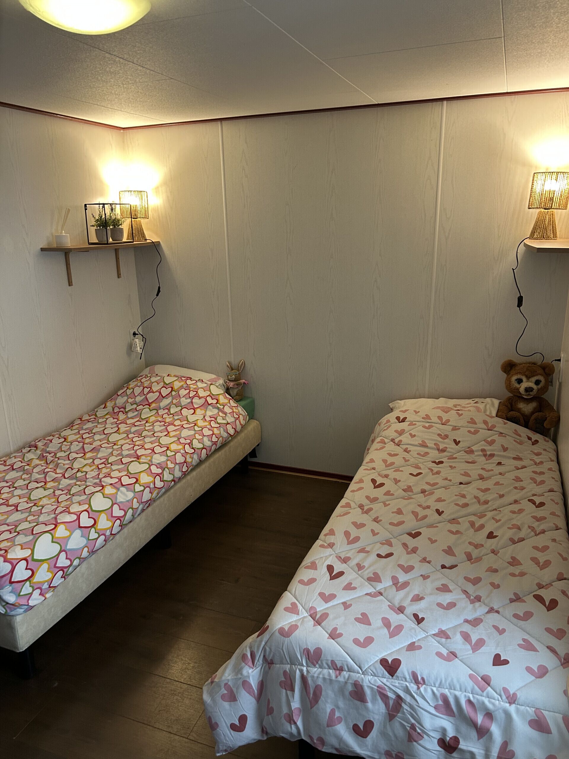 Una stanza con due letti singoli, ognuno con copripiumino a fantasia cuore, un piccolo ripiano con una lampada e un orsacchiotto sul letto a destra.