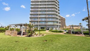 A multi-story hotel with balconies, a garden area with seating, and a clear sky. - 9th Floor Beachfront Condo w/ Pool, Hot Tub, Sauna, & Beach Access! (South Padre Island)
