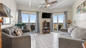 A living room with two sofas, a ceiling fan, a bookshelf, and a TV. - 9th Floor Beachfront Condo w/ Pool, Hot Tub, Sauna, & Beach Access! (South Padre Island)