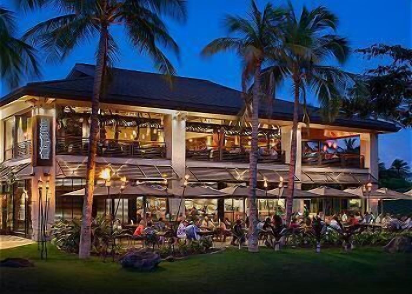 Un hotel de varios pisos con mesas al aire libre y palmeras.