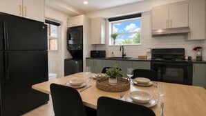 A modern kitchen with a dining table set for two, a black refrigerator, a microwave, a stove, and a window with a view of greenery. - Boho Chic, 3 Bedrooms + Parking + AC | U8 (Québec City)