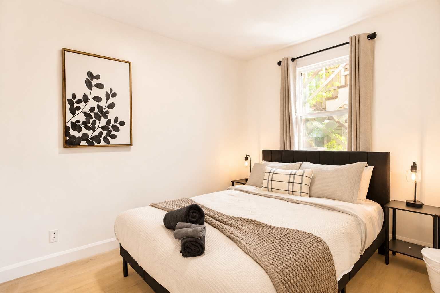 A bedroom with a bed, a nightstand, a wall-mounted light, a window with curtains, and a framed artwork on the wall.