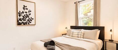 A bedroom with a bed, a nightstand, a wall-mounted light, a window with curtains, and a framed artwork on the wall.