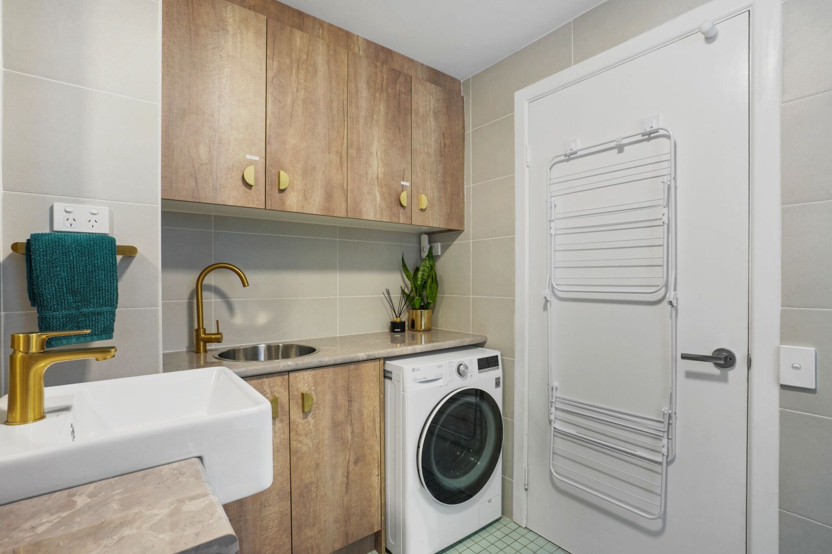 A compact kitchen with a washing machine, a sink with a gold faucet, wooden cabinets, and a towel rack.