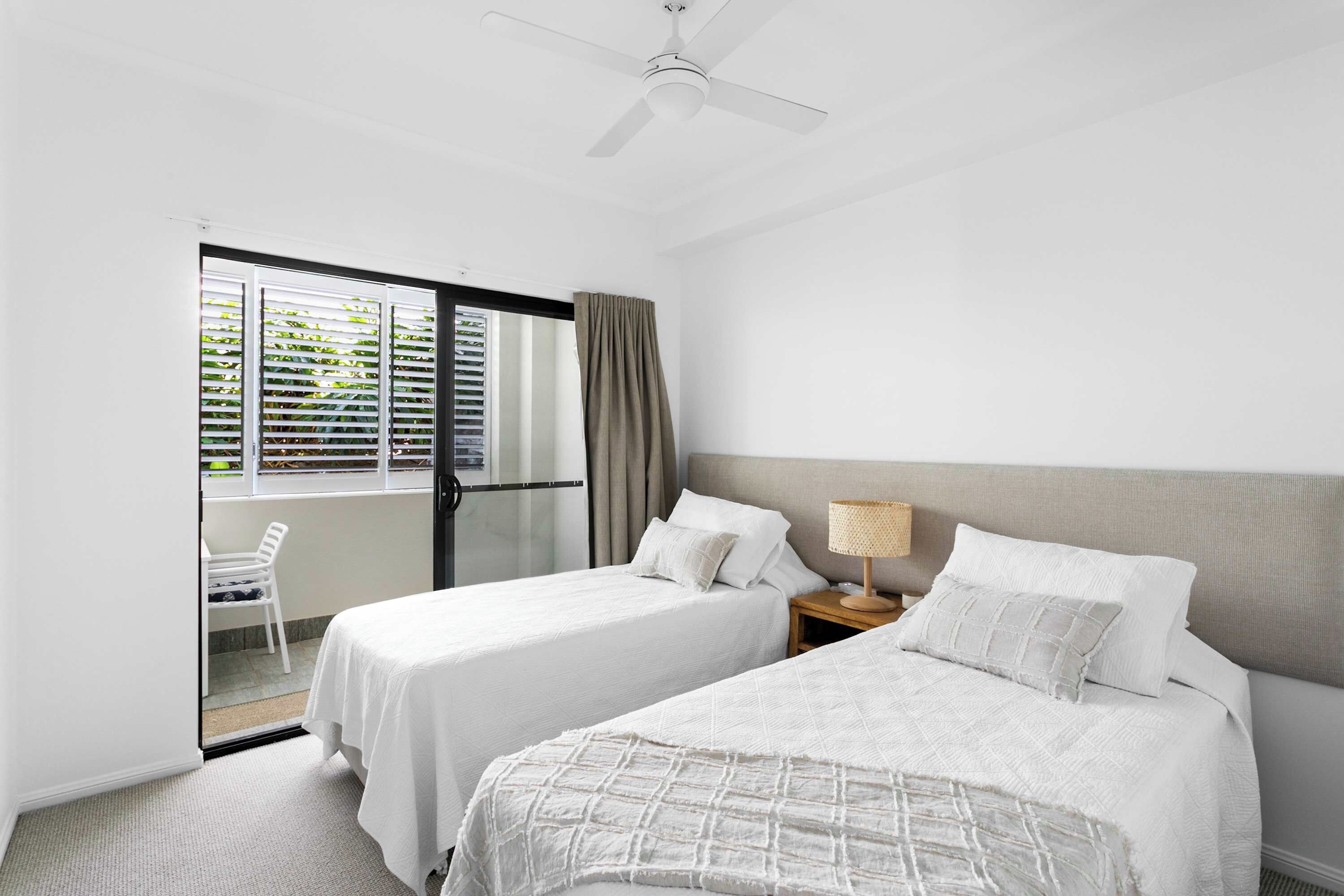 A hotel room with two beds, a ceiling fan, a small table with a lamp, and a sliding glass door leading to a balcony.