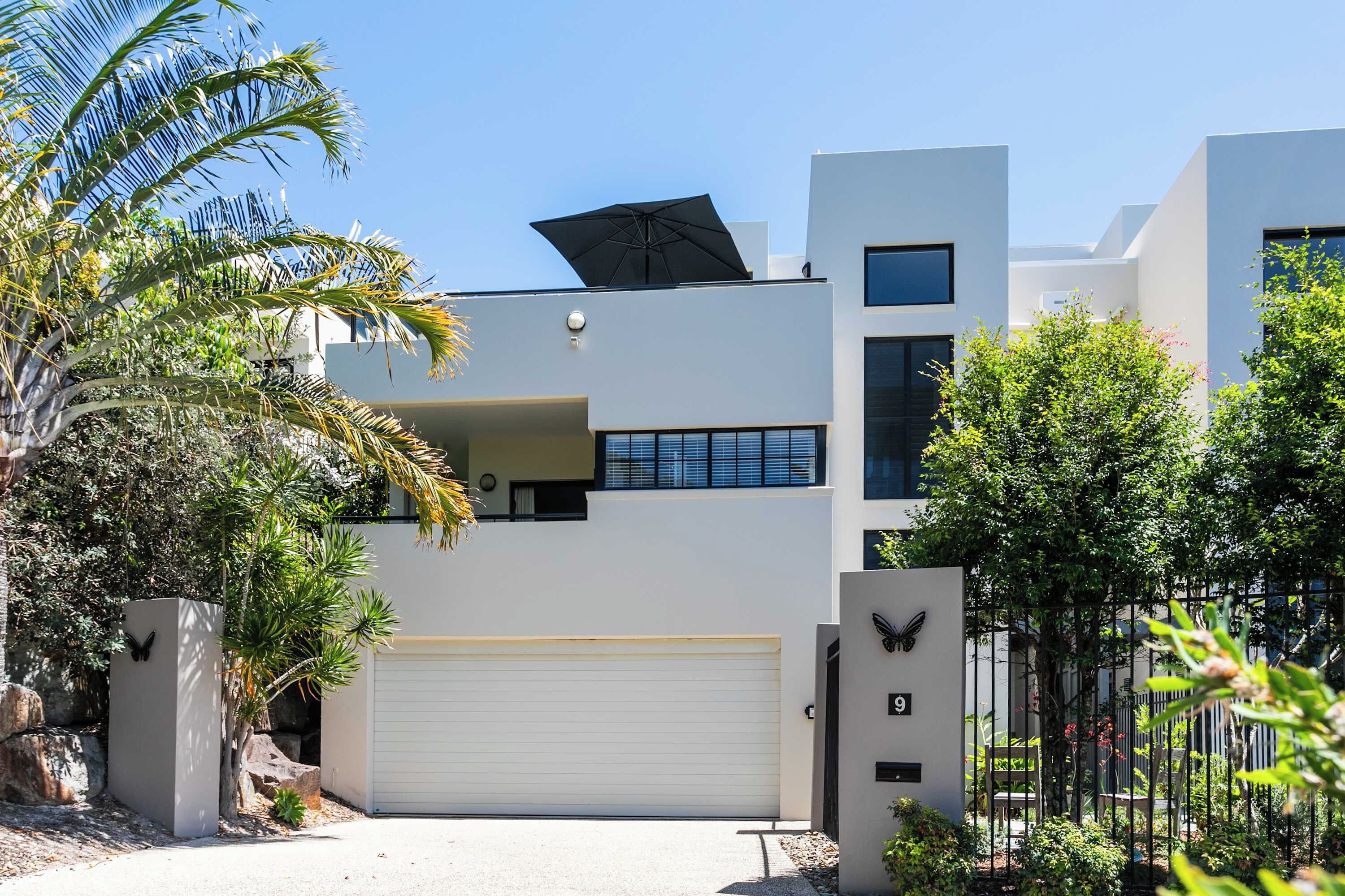 Une maison moderne à deux étages avec un garage, entourée de plantes tropicales et d’une clôture en métal.