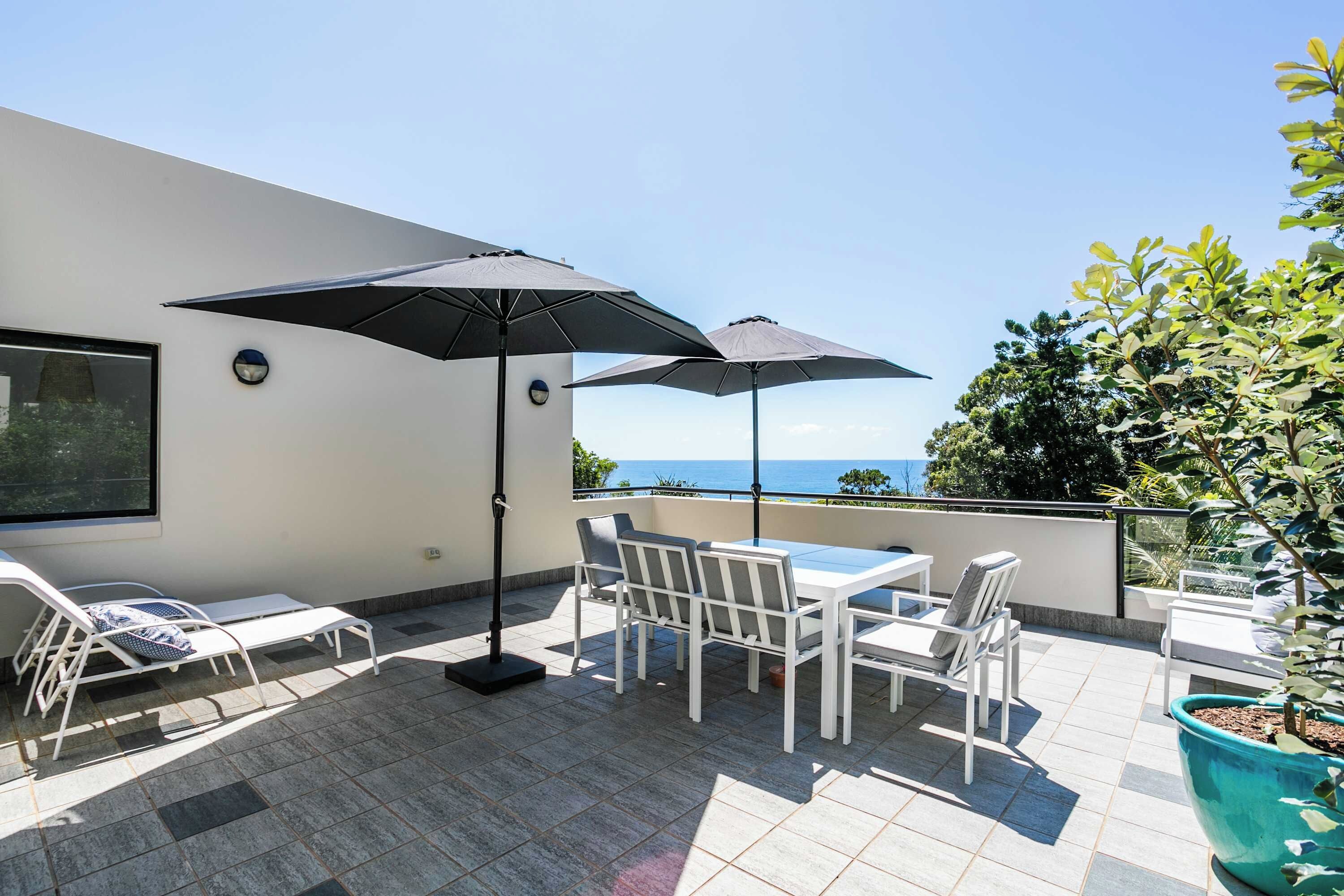 A terrace with a table and chairs, two umbrellas, and a view of the sea.