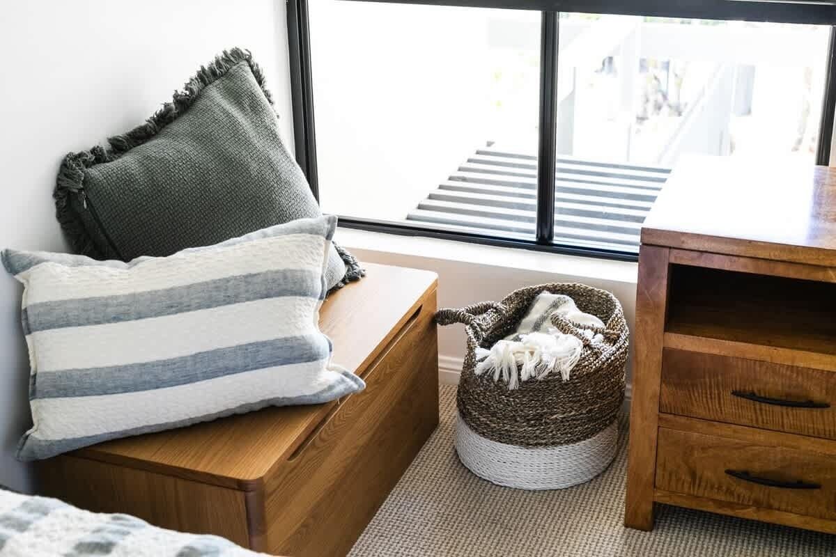 A room with a wooden bench, a striped pillow, a woven basket with a white blanket, and a view of a staircase through a window.