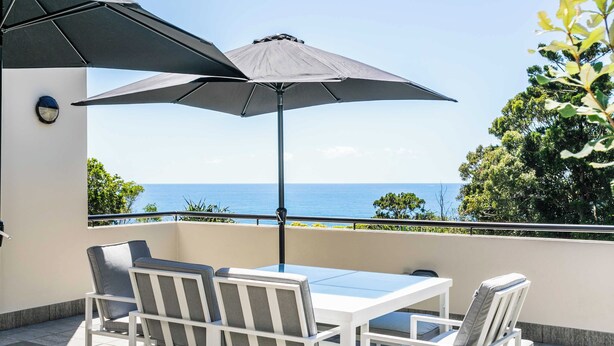 Un balcon avec une table et des chaises, un grand parasol et une vue sur la mer.