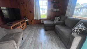 A living room with a television, a sofa, a chair, a small table, and a window with curtains. - 🐦 Wren’s Place 2B/1Ba | Charming Seaside Retreat • Walk to Beach • Firepit (Seaside)