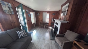 A cozy living room with a fireplace, wooden walls, and a dining area. - 🐦 Wren’s Place 2B/1Ba | Charming Seaside Retreat • Walk to Beach • Firepit (Seaside)