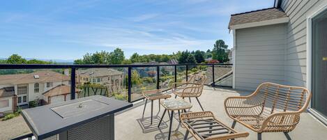Een balkon met rieten meubilair, een glazen hekwerk en uitzicht op omliggende huizen.