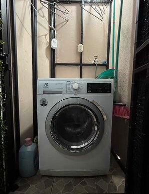 Eine kompakte Waschmaschine in einem kleinen Waschraum mit Fliesenboden, Wasserhahn und Steckdose.