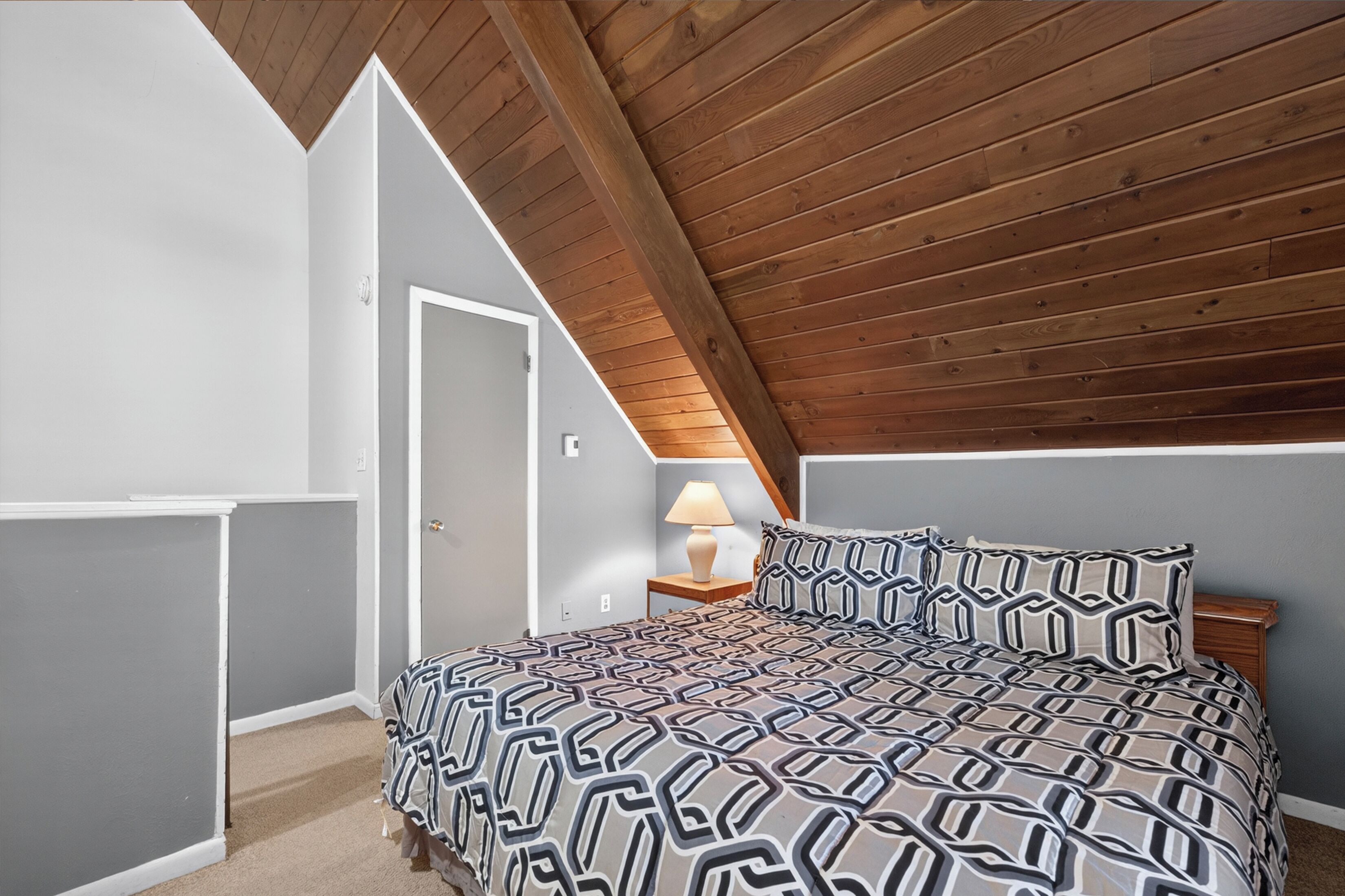 Un dormitorio con techo inclinado, una cama con ropa de cama estampada, una mesita de noche con una lámpara y una puerta.