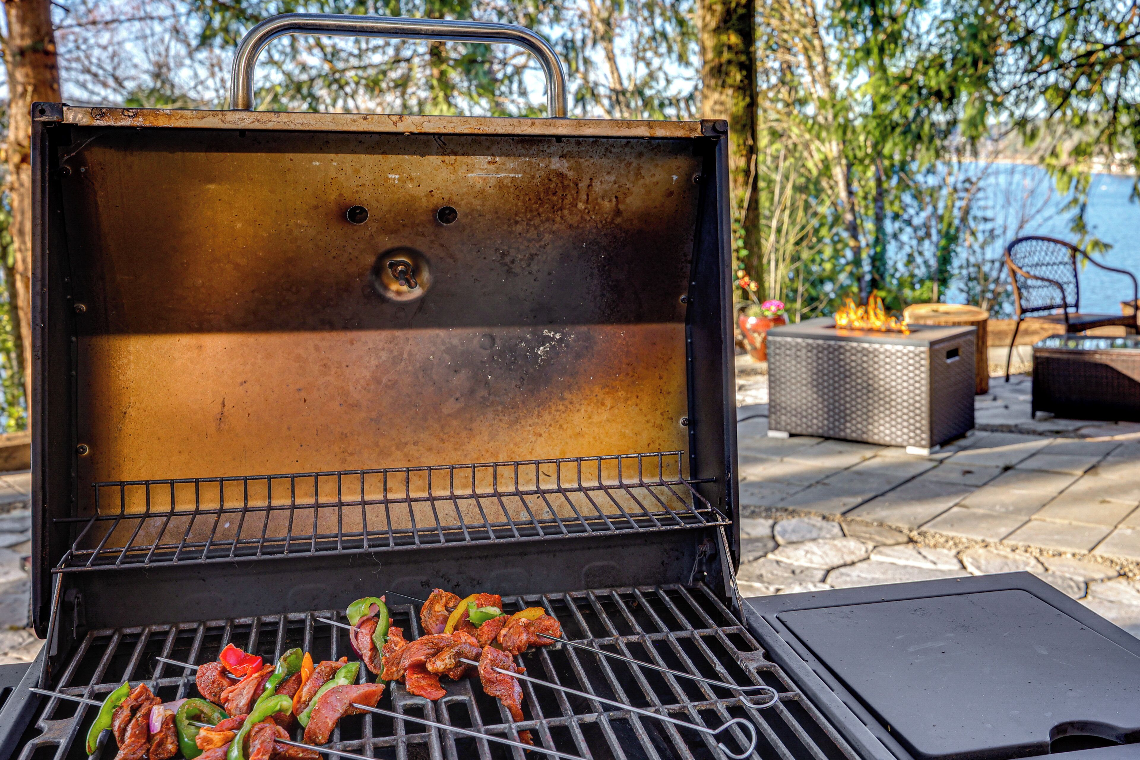 Ein Grill mit Fleisch- und Gemüserspießen, einer Metall-Grillabdeckung und Gartenmöbeln.
