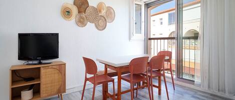 A room with a TV on a wooden cabinet, a table with four chairs, and a balcony with a view of buildings.