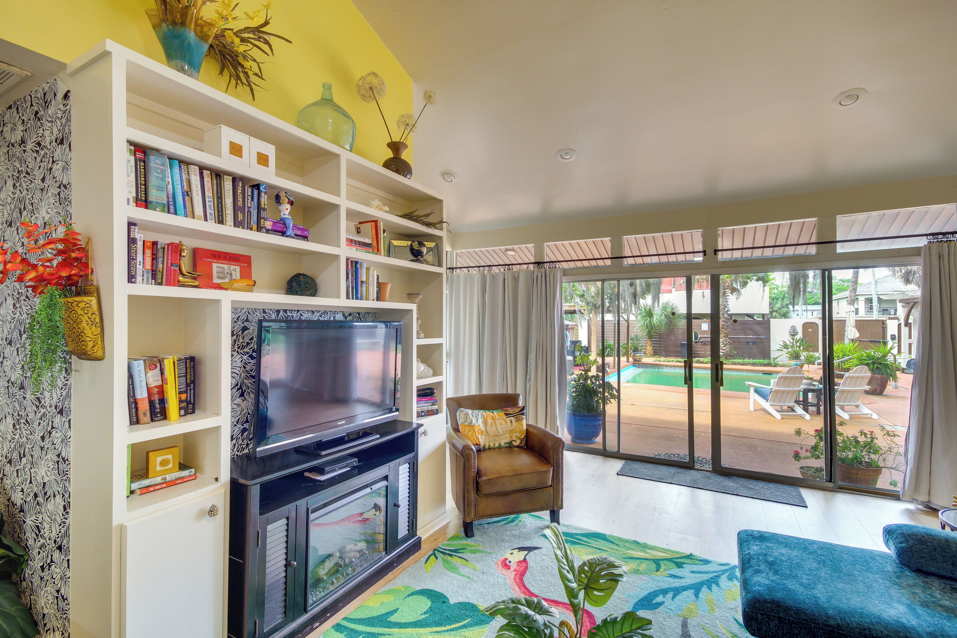 Ein Wohnzimmer mit Fernseher, Bücherregalen, einer Couch und Blick auf einen Pool und Sitzgelegenheiten im Freien.