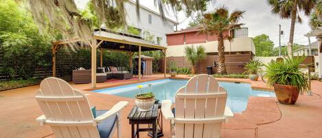 A backyard with a pool, two Adirondack chairs, a covered seating area, and a house in the background.