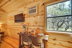 A wooden cabin interior with a dining area, a window with a view of trees, and a wall-mounted air vent. - 2 Studio Cabins! Hill Country Escape w/ Fire Pit (Johnson City)