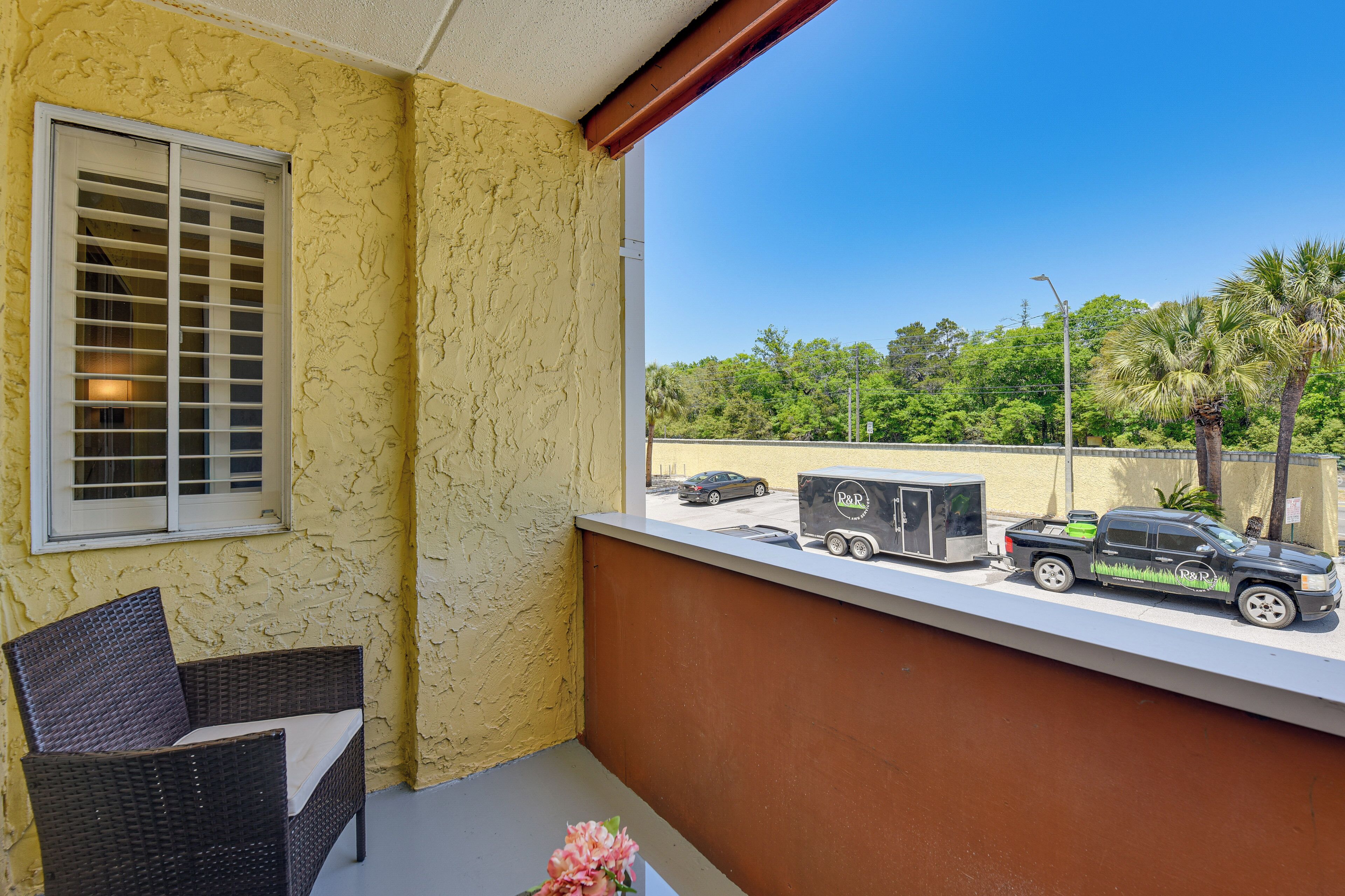 A balcony with a wicker chair, a flower, and a view of parked vehicles and greenery.