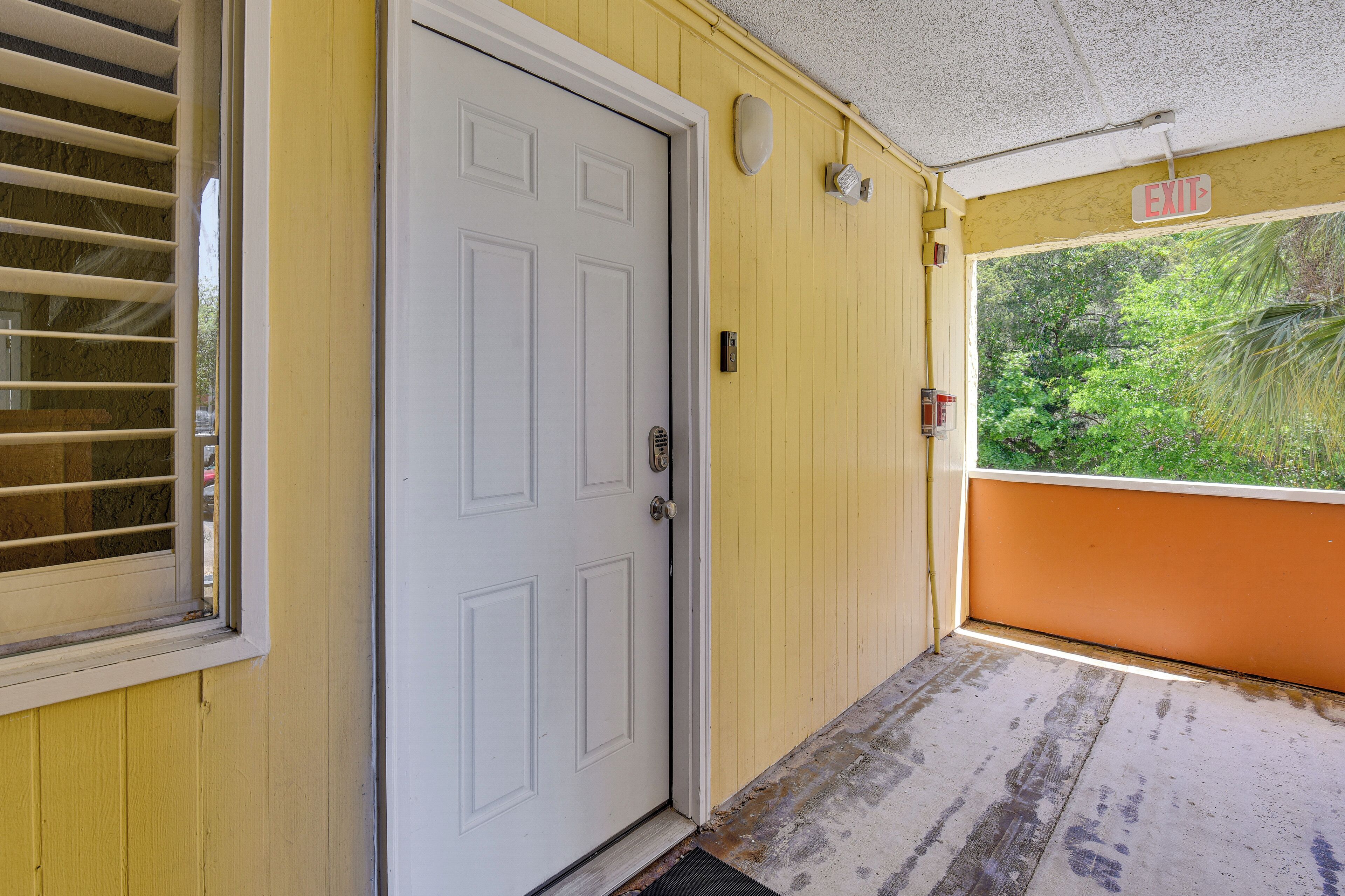 A yellow exterior wall, a white door, a window with blinds, and a view of greenery.