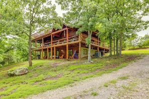 A rustic cabin with a wooden deck and multiple levels, surrounded by lush greenery. - Game Room + Roof Deck: Ozarks Group Getaway! (Harrison)