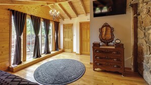 A room with wooden beams, a chandelier, a large window with curtains, a wooden dresser, and a flat-screen TV mounted on a stone wall. - Game Room + Roof Deck: Ozarks Group Getaway! (Harrison)