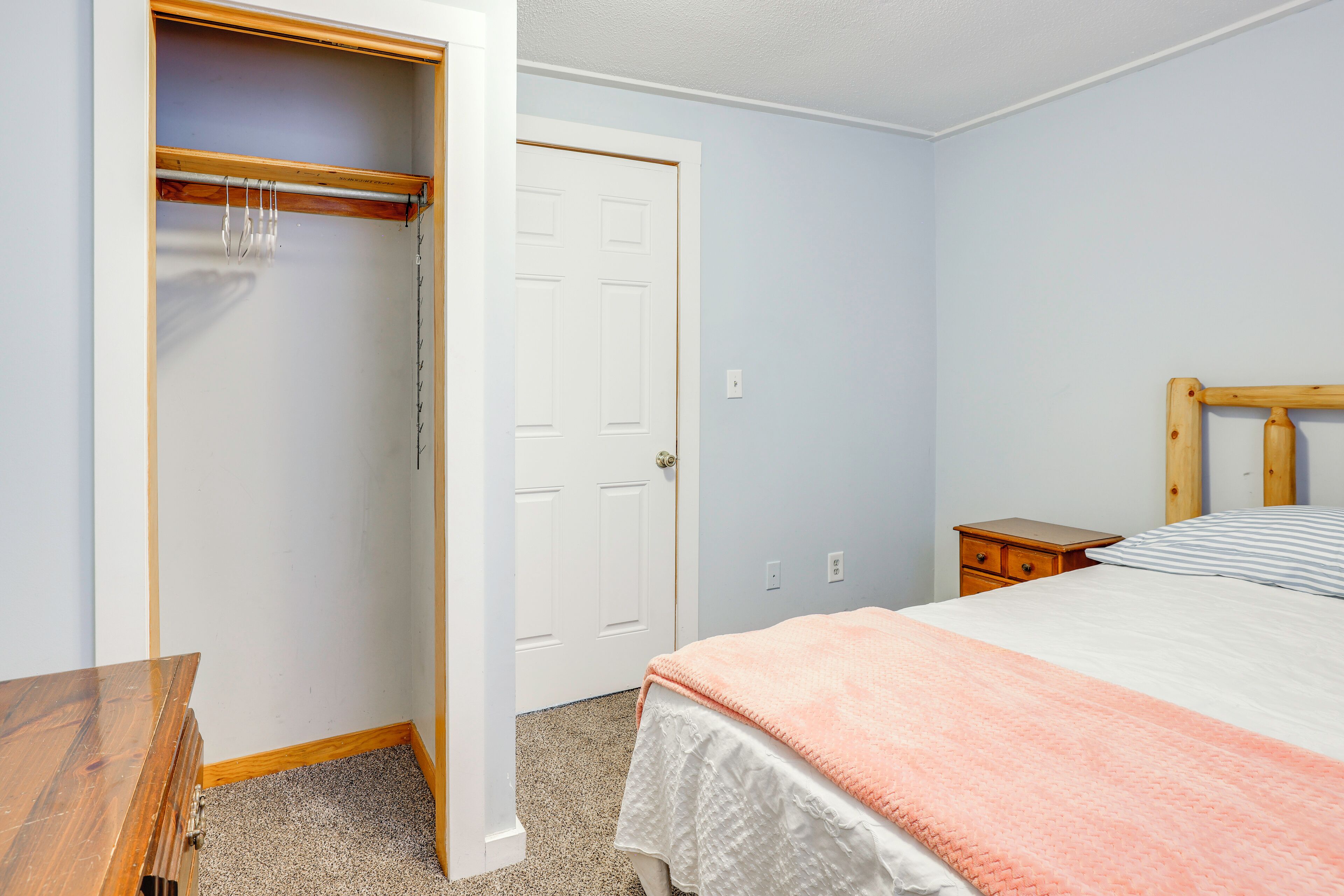 A bedroom with a bed, a wooden nightstand, a wooden dresser, a closet with hanging clothes, and a white door.