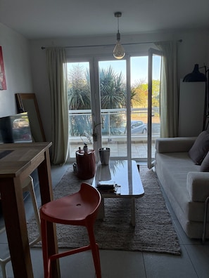 Un salon avec un canapé, un fauteuil rouge, une table basse et une table à manger. Il y a une grande fenêtre avec des rideaux, et de là, on peut voir des arbres et un parking.