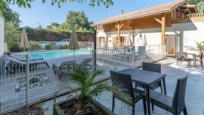 Uma área de piscina com espreguiçadeiras e guarda-sóis, um pátio cercado com mesas e cadeiras, e um prédio com fachada de madeira e varanda.