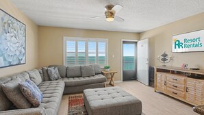 Ein Wohnzimmer mit Ecksofa, Hocker, Holzschrank, Deckenventilator und einem großen Fenster mit Jalousien.