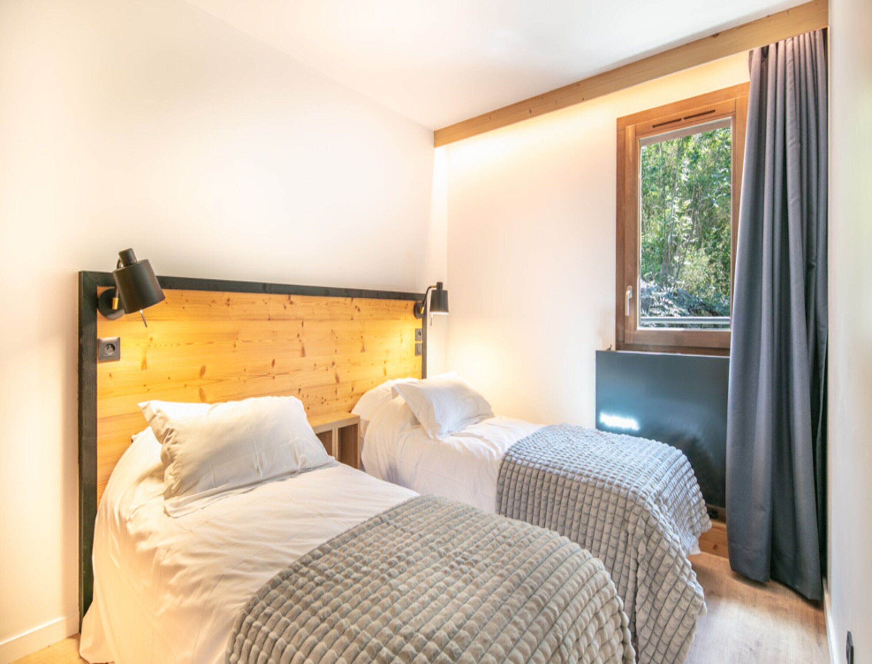 Une chambre avec deux lits, une tête de lit en bois, une fenêtre avec des rideaux et une vue sur la verdure.