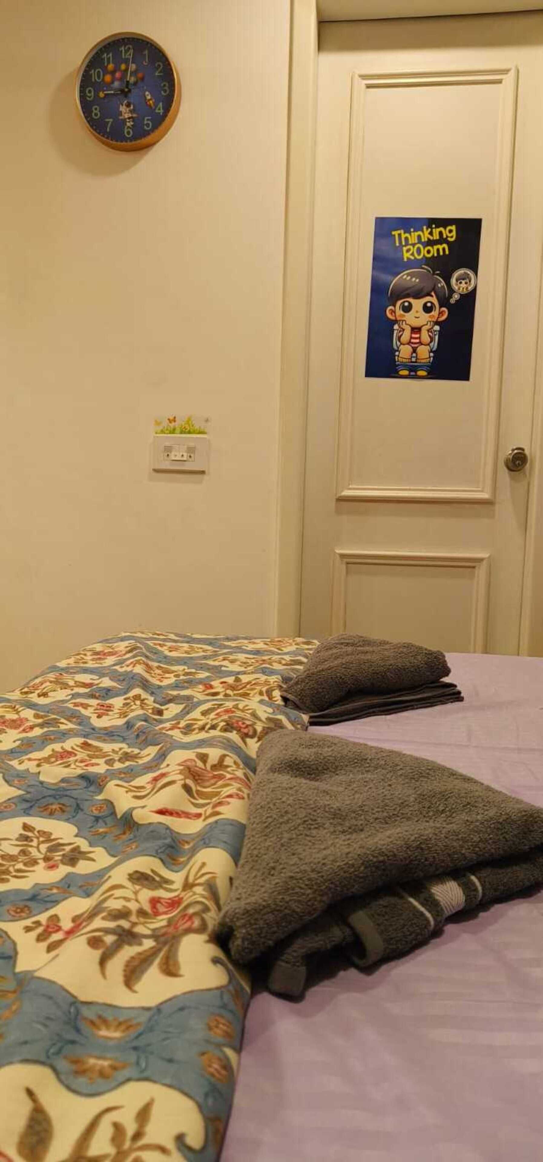 A bedroom with a bed, a wall clock, a door with a 'Thinking Room' sign, and a power outlet.