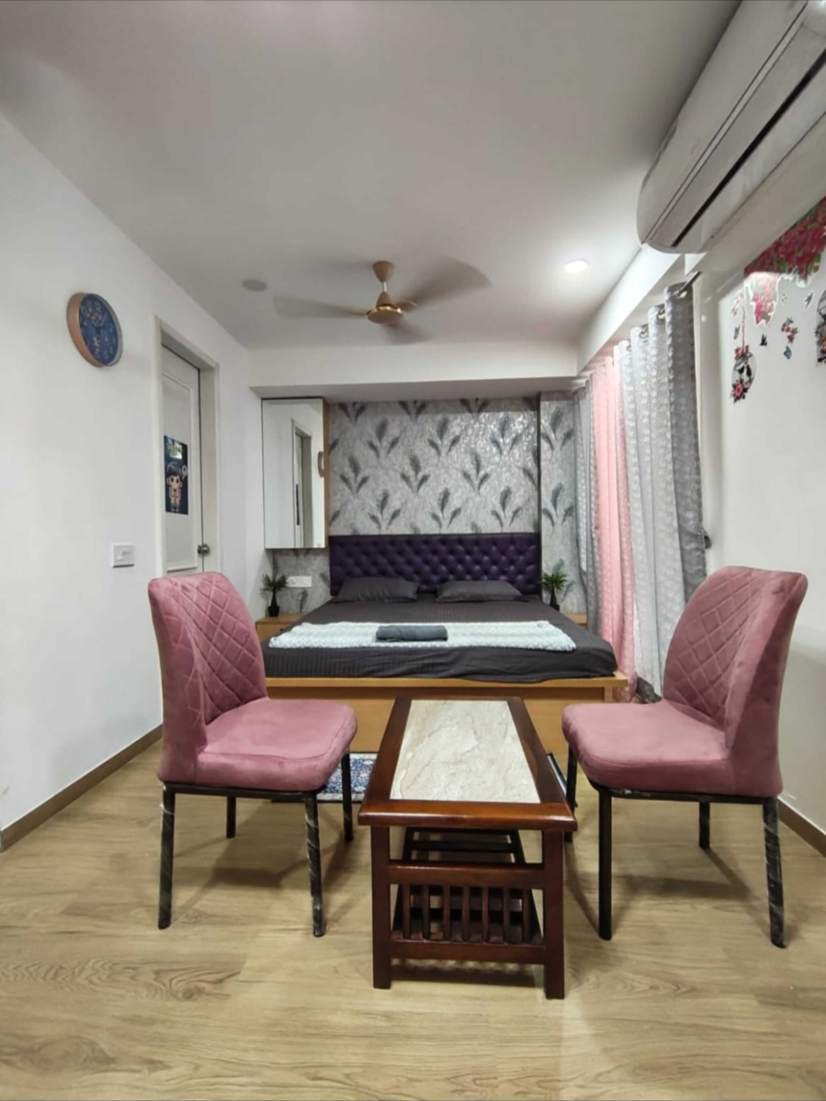 A bedroom with a bed, two chairs, a small table, an air conditioner, and a ceiling fan.