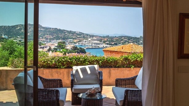 A balcony with wicker furniture and a glass door overlooking a coastal town. - Villa Hermes - Porto Cervo Center (Porto Cervo)