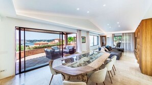 A modern interior with a glass-top dining table, sofas, and a large window offering a scenic view. - Villa Hermes - Porto Cervo Center (Porto Cervo)