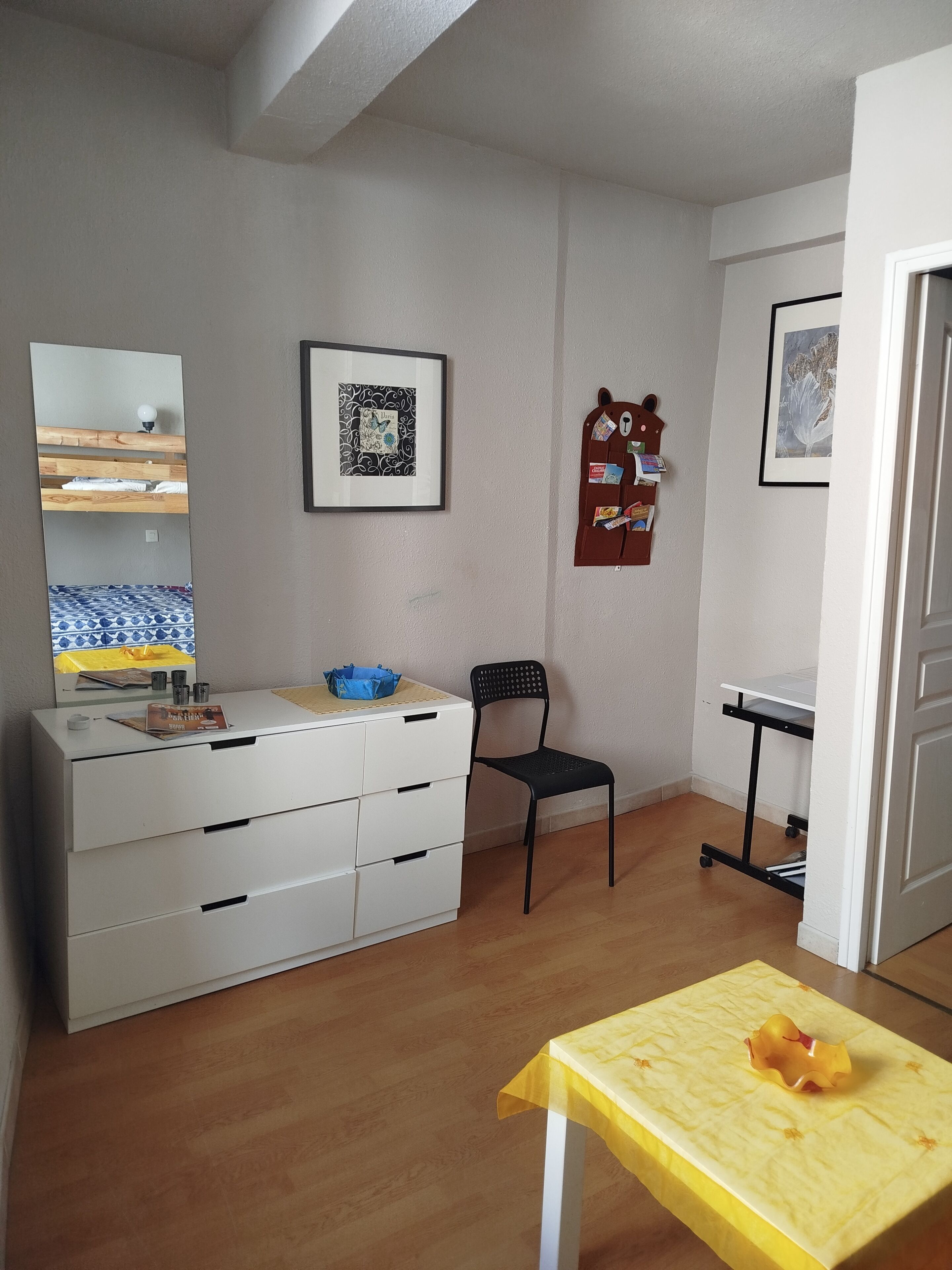A room with a white dresser, a chair, a small table with a yellow tablecloth, a wooden floor, a wall-mounted picture frame, and a door.