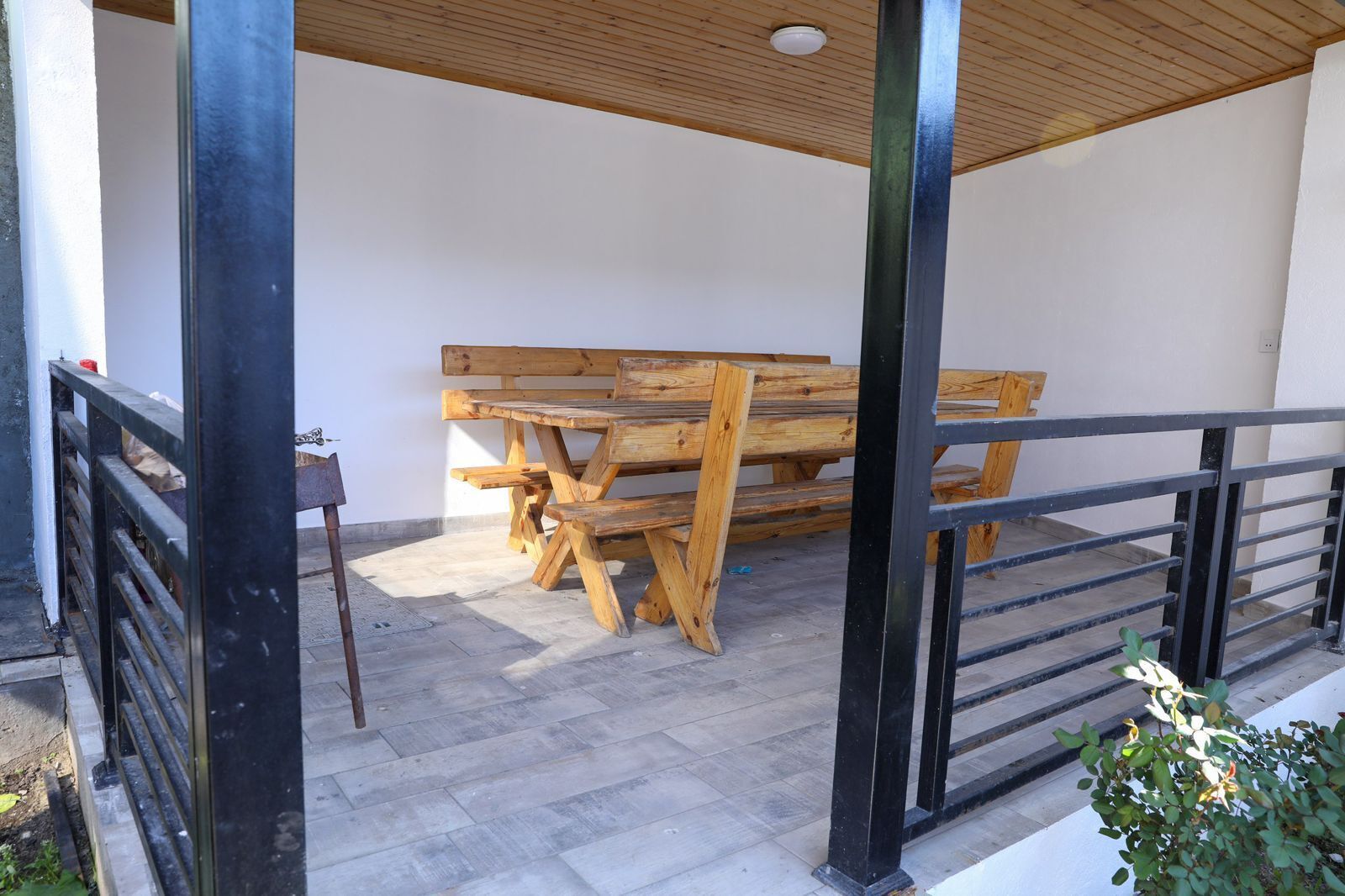 A covered outdoor seating area with a wooden table and benches, a metal railing, and a concrete floor.