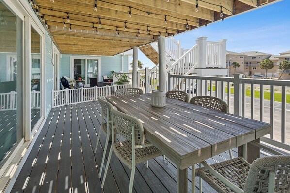 Een overdekt terras buiten met een houten tafel en rieten stoelen, een witte balustrade en uitzicht op de omliggende gebouwen.
