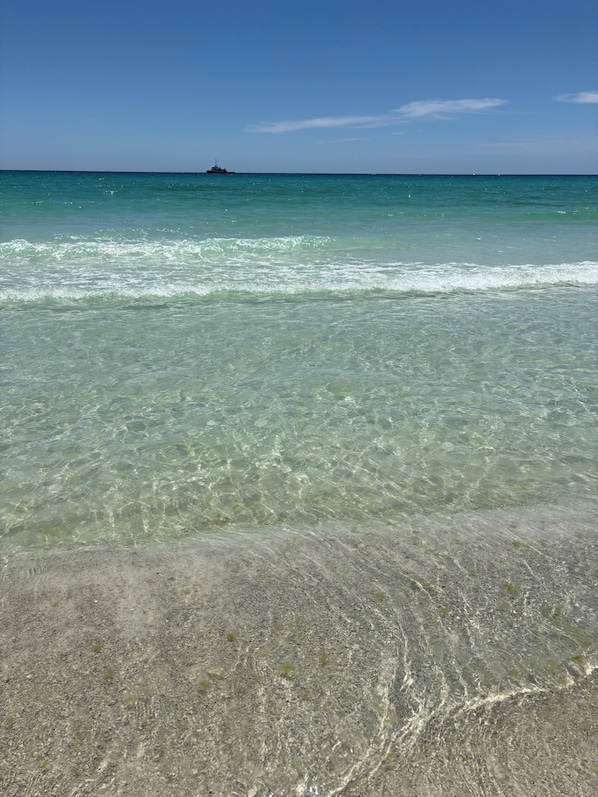 Une plage avec une eau claire et peu profonde, un bateau au loin et un premier plan sablonneux.