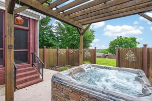 A hot tub under a wooden pergola with steps leading to a door, surrounded by a wooden fence and greenery.