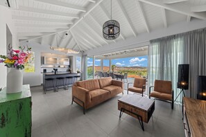 Ein großzügiger Wohnbereich mit einer Couch, einem Couchtisch und Sesseln, eine Küche mit Barhocker und große Fenster mit Blick auf die Landschaft.