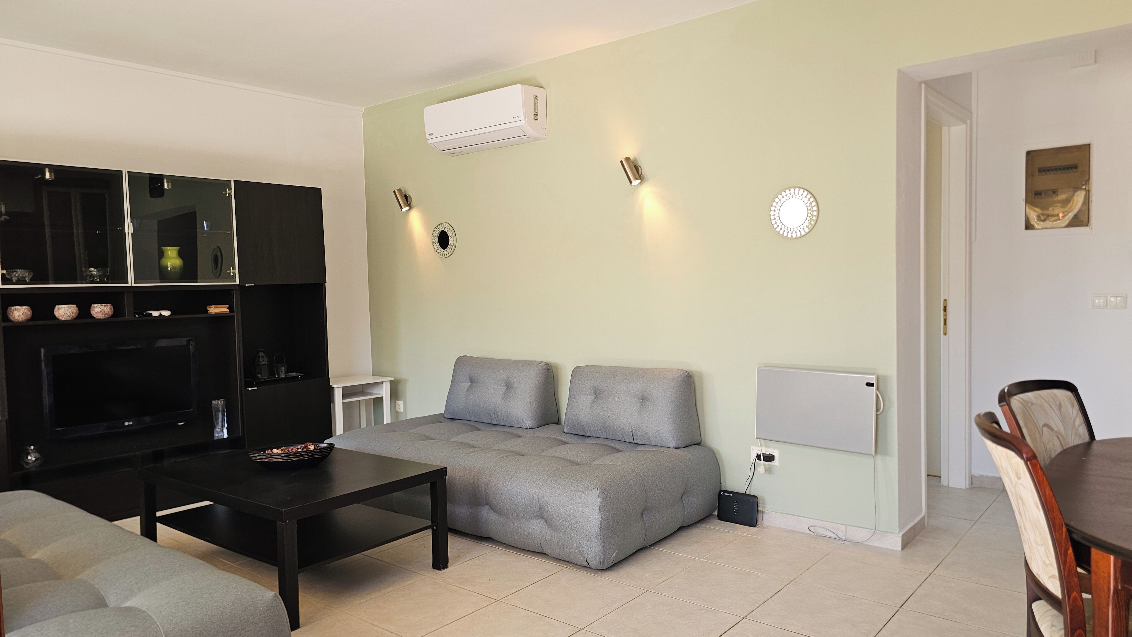 A living room with a grey sofa, black coffee table, and a TV on a black stand. There is a white air conditioner on the wall and a small white table.