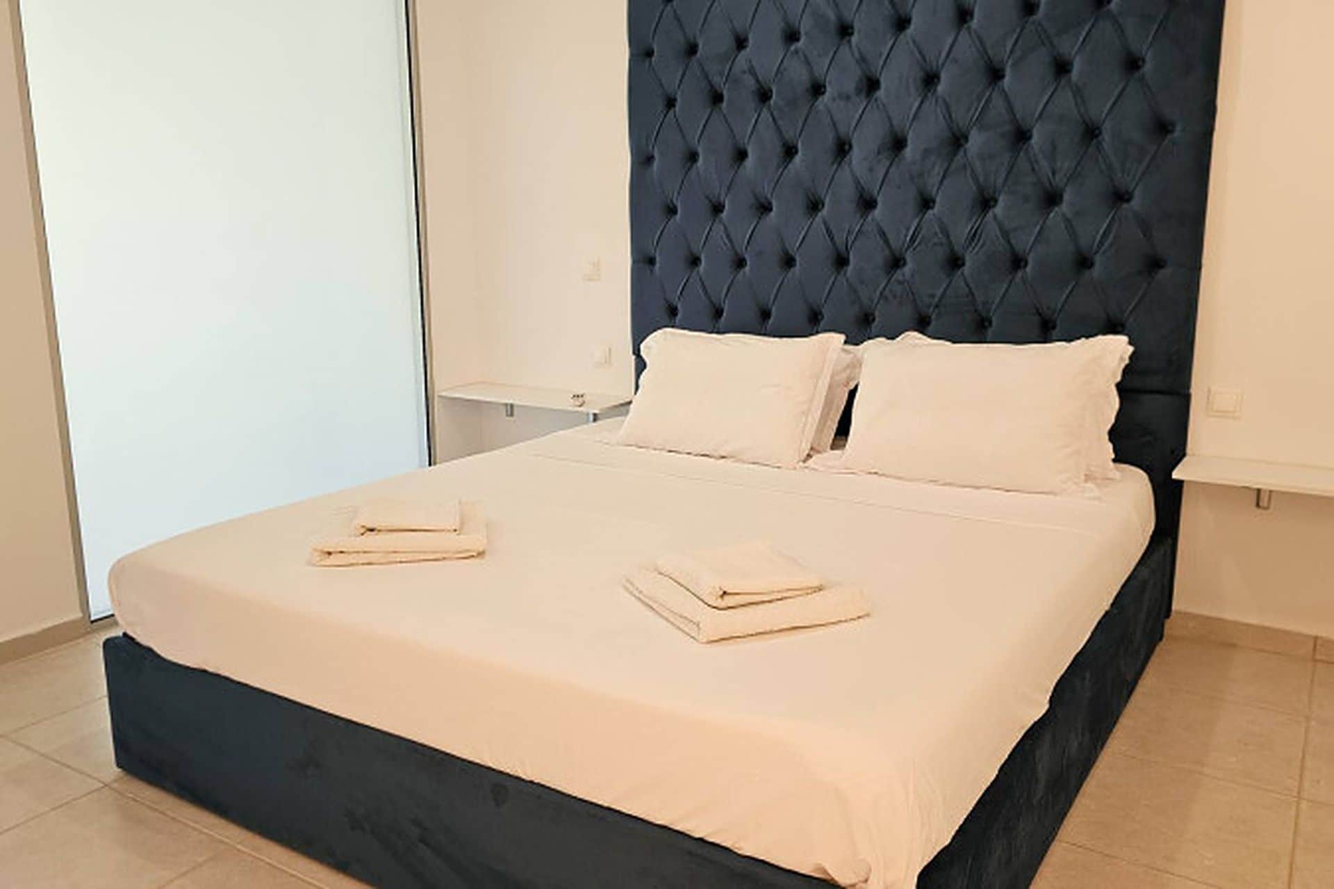 Een bed met een donker hoofdbord en twee kussens, een opgevouwen handdoek op het bed, en een witte muur op de achtergrond.