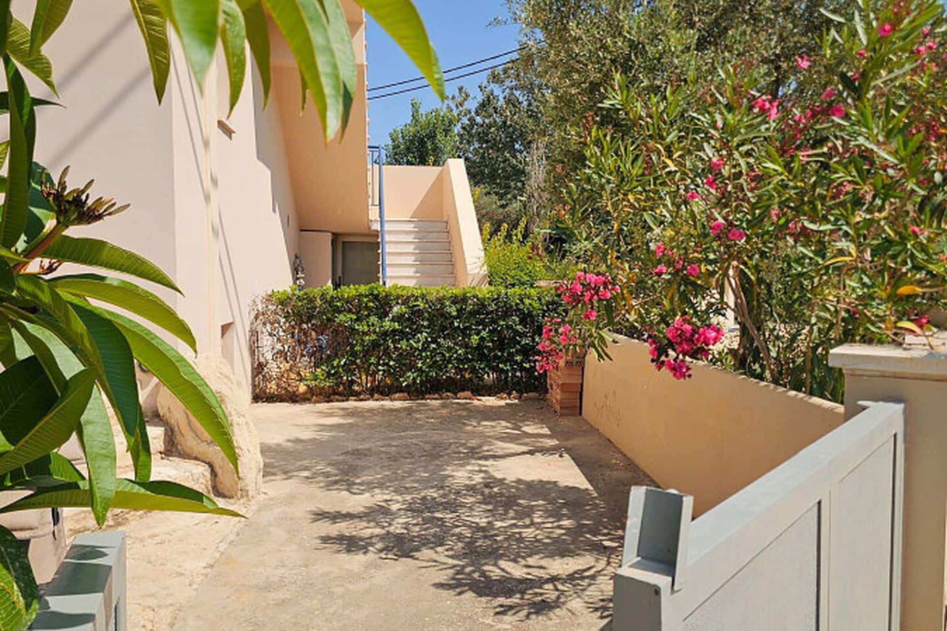 Un sendero estrecho que lleva a un edificio de fachada color beige, rodeado de follaje y plantas con flores.